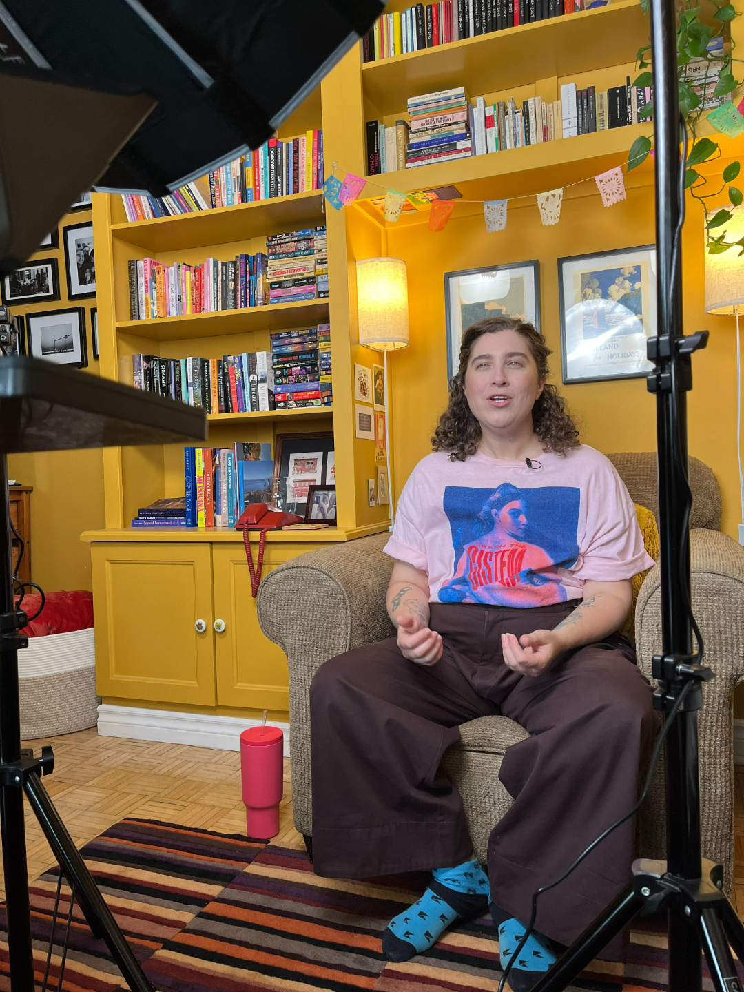Ren&eacute;e sitting in their armchair in front of their bookshelf, being filmed for YouTube