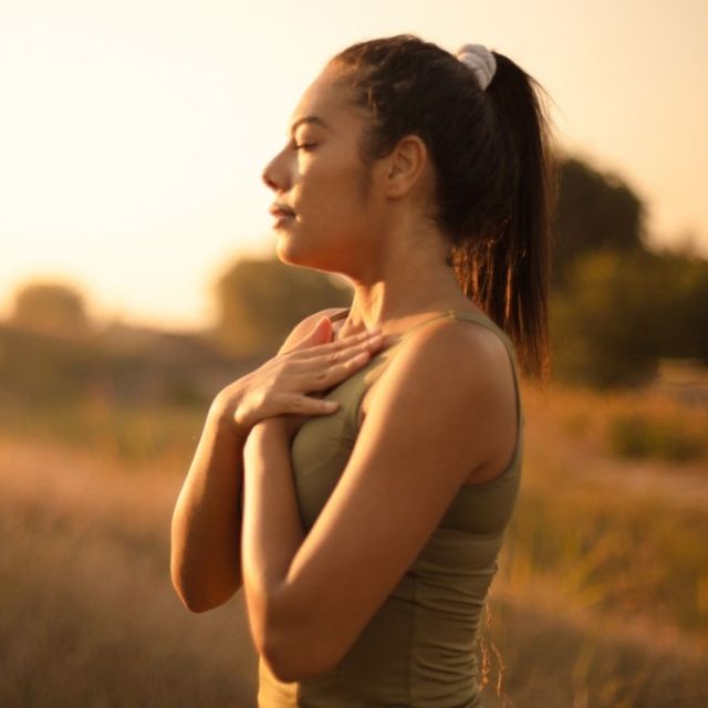 A person in a tank top with eyes closed and hands crossed over their chest, standing in a field at sunset. Janet Farnsworth Full Body Energy Orgasm Retreat 