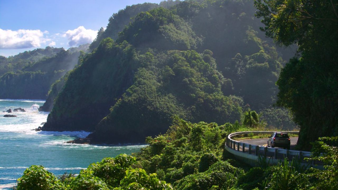 An elevated view of a coastal road Hawaii with a car driving along the guardrail-lined curve, with a lush, green, mountainous coastline in the background.Full Body Energy Orgasm Retreat 