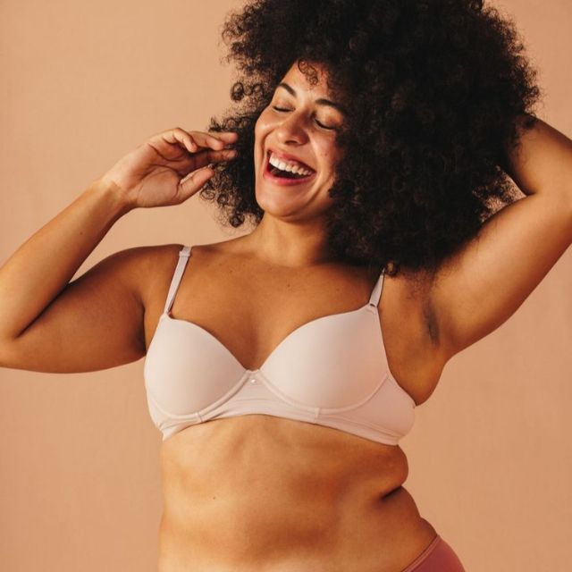 A smiling person with curly hair, wearing a bra and underwear, poses against a peach-colored background.Janet Farnsworth Full Body Energy Orgasm Retreat 