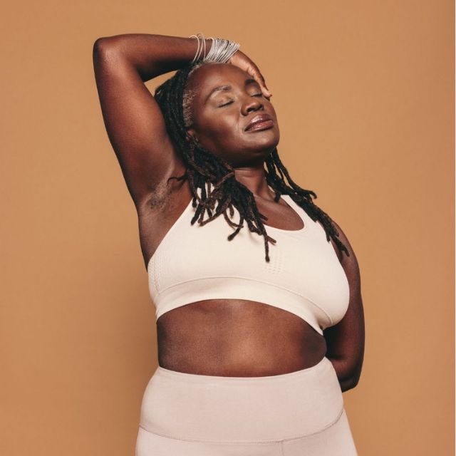 A person with dreadlocks wearing leggings and a sports bra with one arm up and eyes closed, on a brown Janet Farnsworth Full Body Energy Orgasm Retreat background.