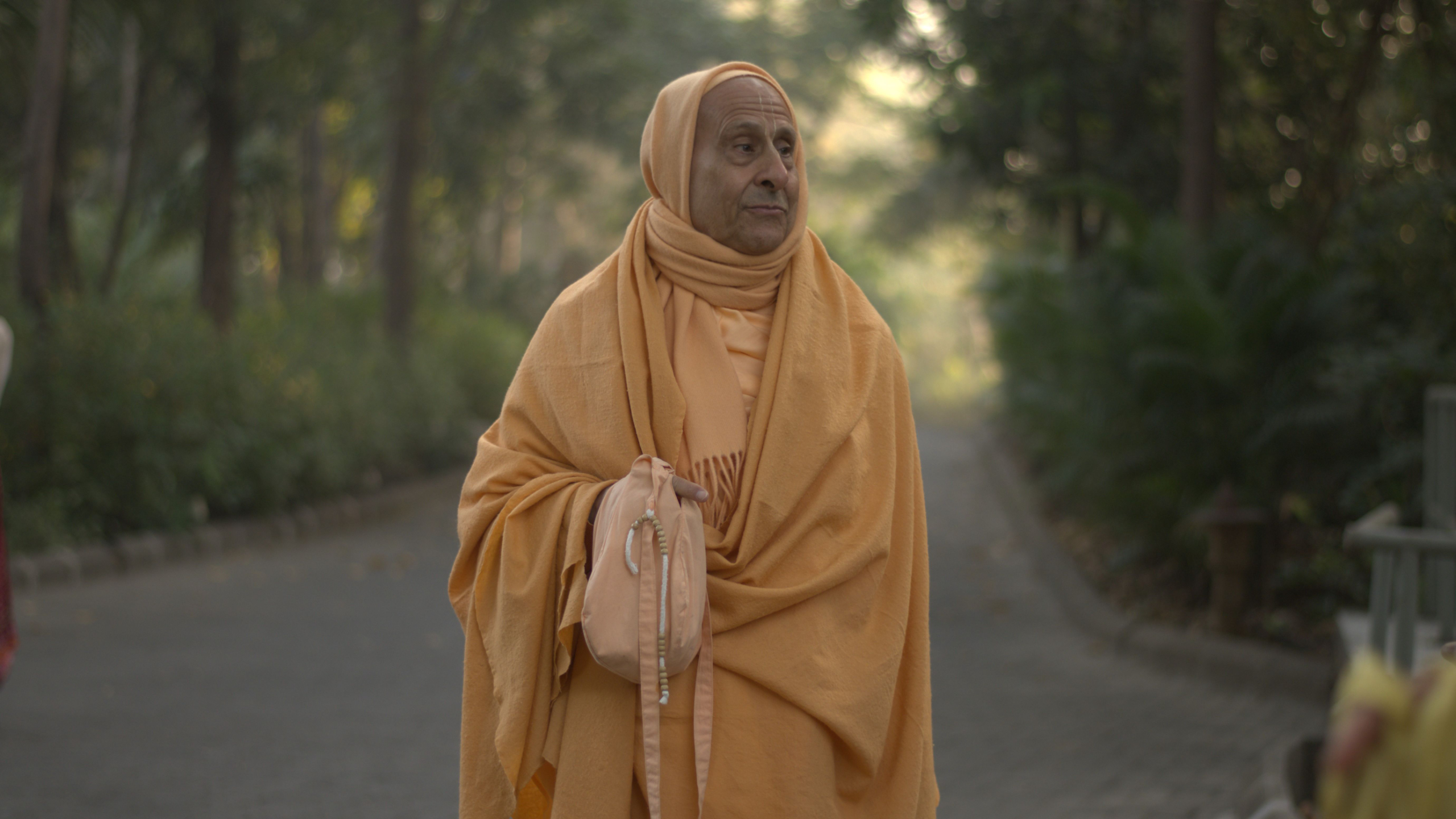 Radhanath Swami in traditional Vaishnava robes walking outdoors on a quiet path surrounded by greenery in India.