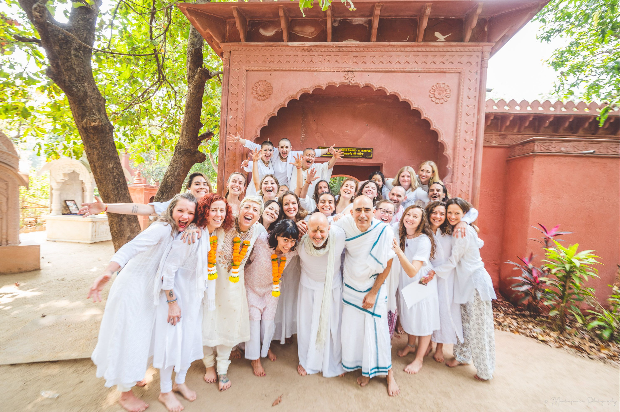 Group of yoga teacher training students and instructors dressed in white, smiling and celebrating together in front of a traditional red sandstone archway at Govardhan Eco Village in India, symbolizing community, devotion, and completion of the training.