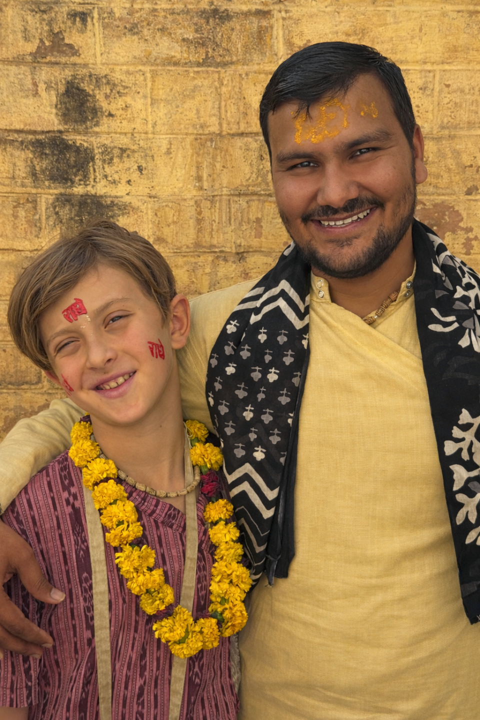 Smiling pilgrim and local devotee in India, wearing traditional clothing, tilak markings, and marigold garlands against a temple wall.