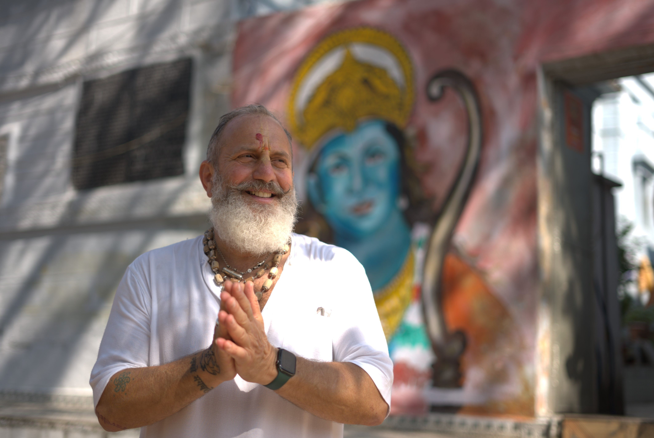 Raghunath Cappo smiling with hands in prayer in India, standing before a devotional mural during a bhakti yoga pilgrimage.