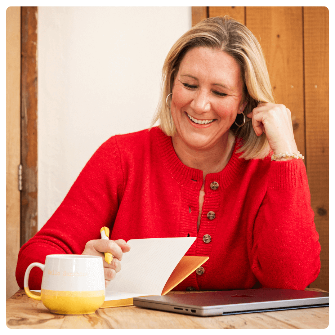 Karen Sutton sitting at a wooden table with a laptop, yellow notebook, and a glass of water, smiling warmly. The background features a teal couch, yellow flowers, and framed pictures, representing her blog on grief support.
