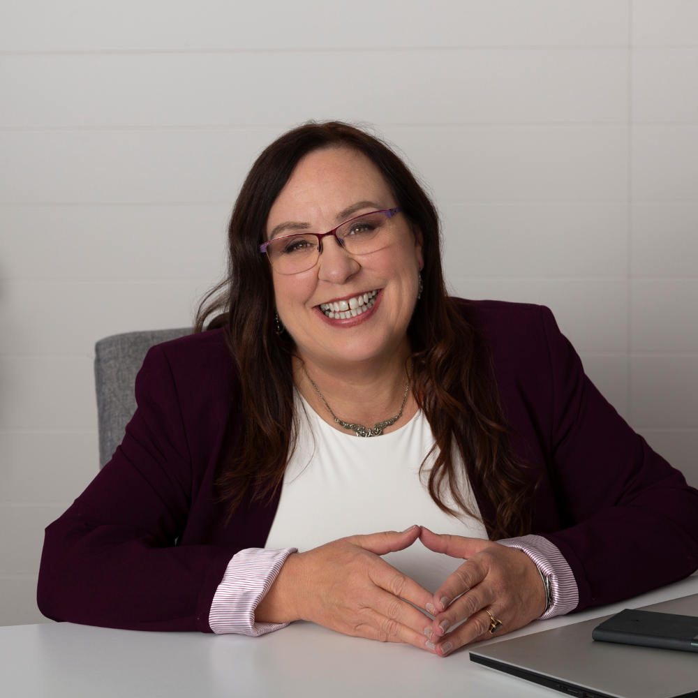 Amanda L. Christin with long brown hair and glasses sits cross-legged on a soft rug, eyes closed and one hand resting over her heart in a peaceful, reflective pose. She wears a charcoal-gray top and deep plum pants, surrounded by dark, feathery textures that frame the scene.