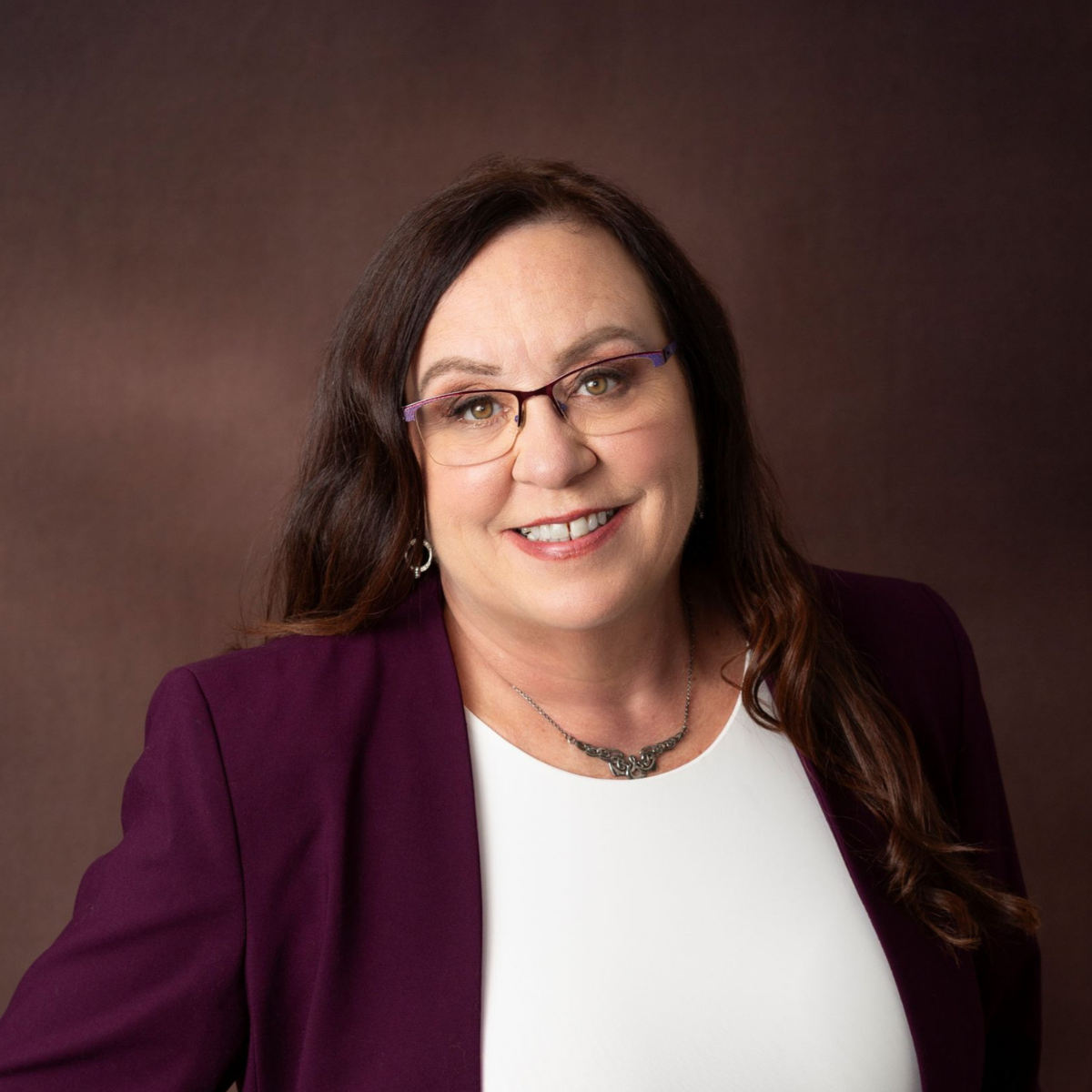 Amanda L. Christian wearing a deep plum blazer and white top, smiling confidently toward the camera in front of a soft neutral background — reflecting warmth, approachability, and executive-level leadership presence.