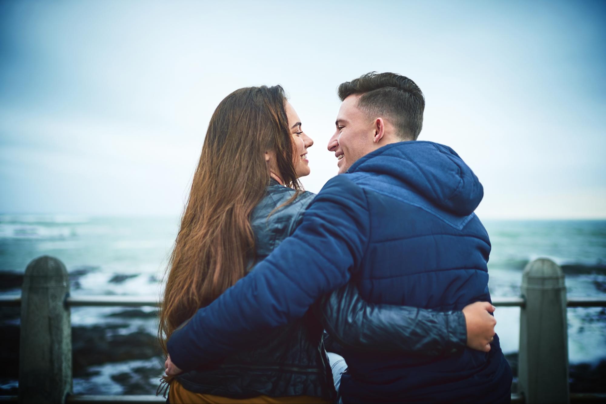 A young couple embrace at a seafront railing, smiling as they look at each other
