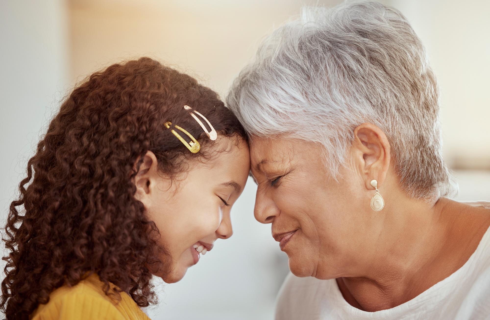 A grandmother and her granddaughter press their foreheads together, both smiling with their eyes closed
