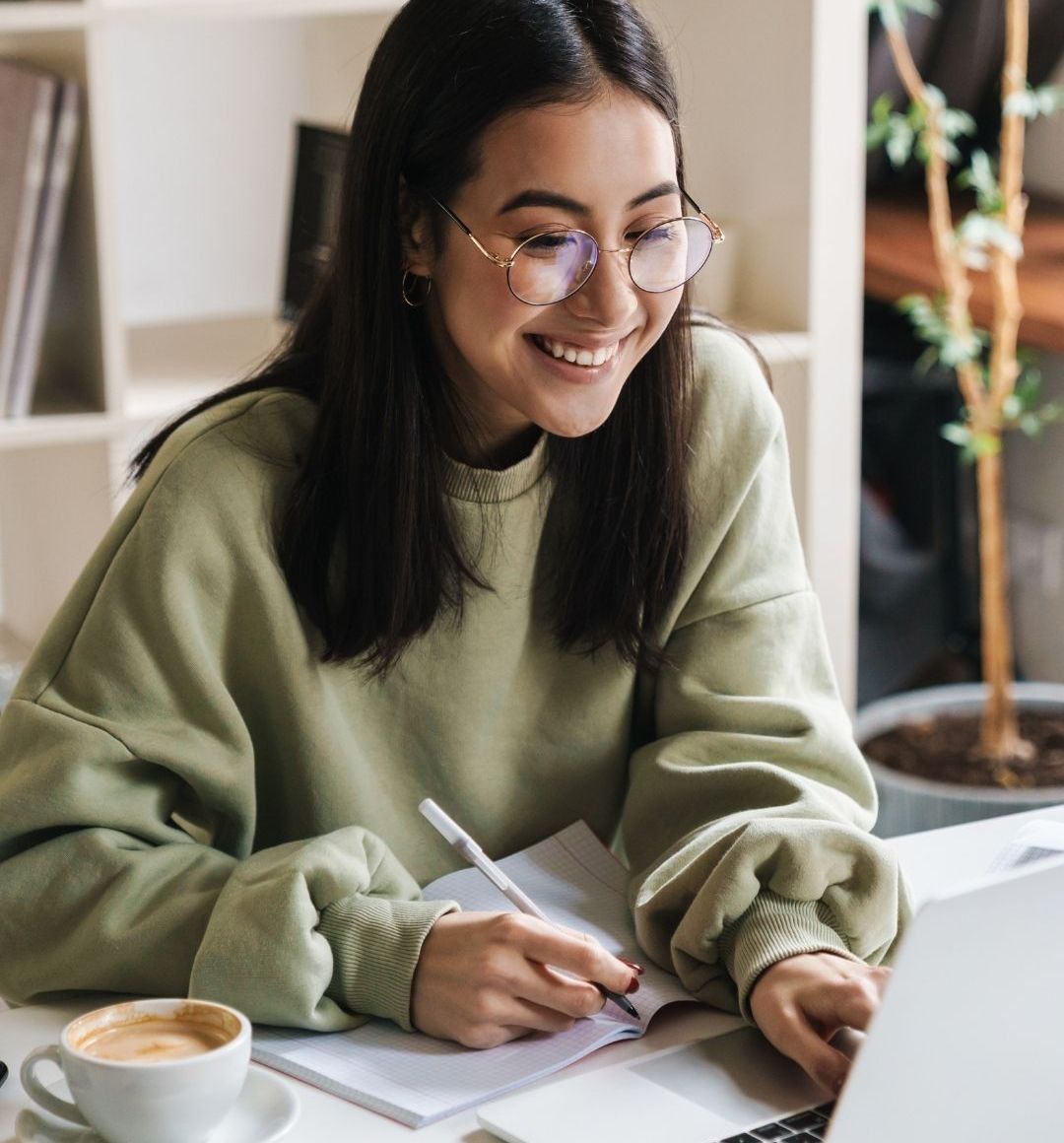 A student smiling while taking notes beside a laptop and coffee, showing how a clear and supportive learning experience can make studying feel calm and manageable rather than overwhelming.