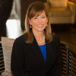 Suzanne Ogle smiling in a patterned jacket and pink blouse sitting by a large window.