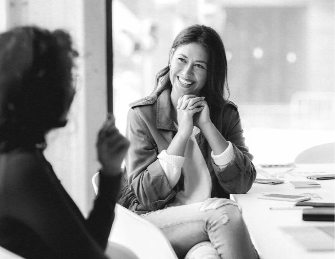 woman sat at a table smiling with her hands together chatting to a friend