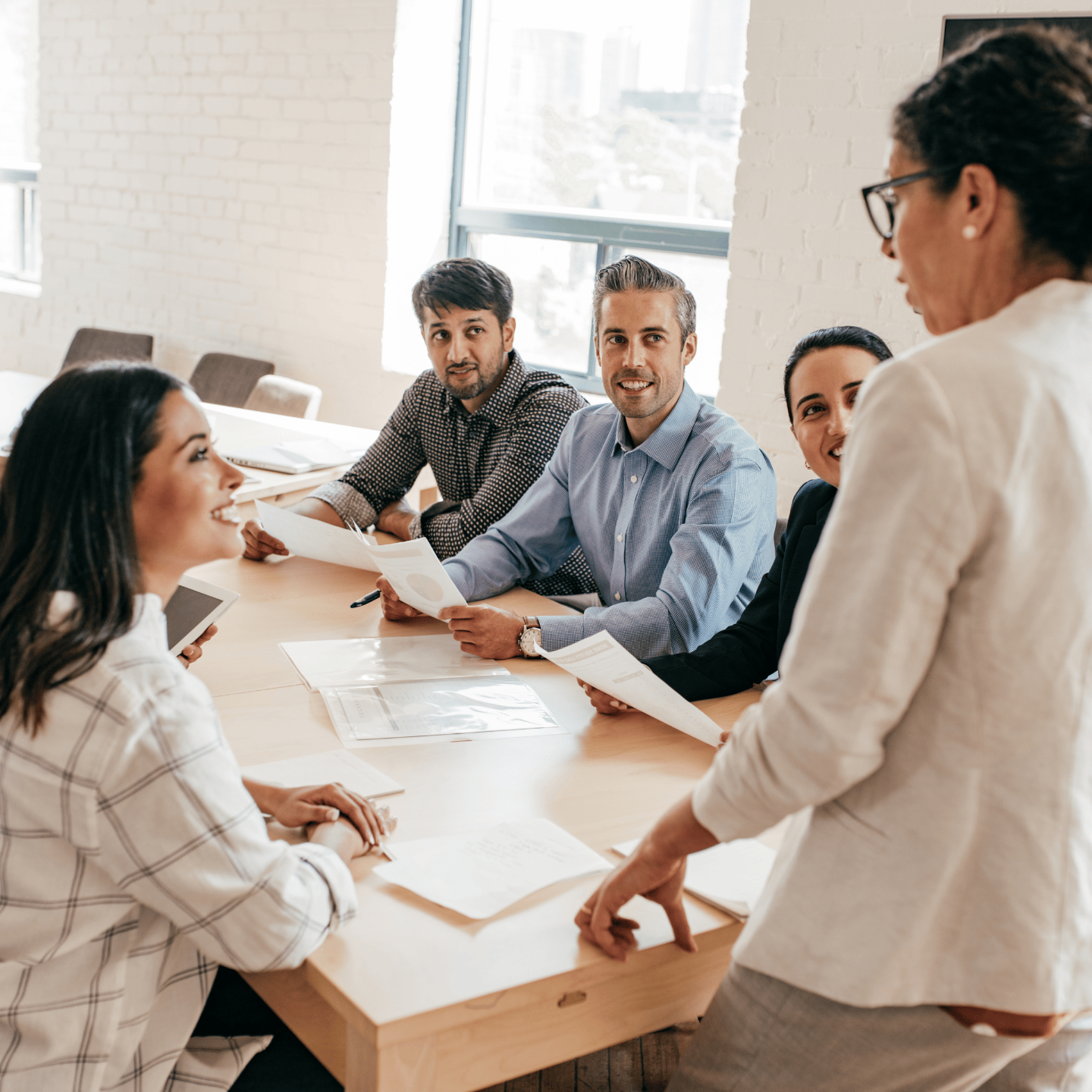 A woman wearing glasses standing at a head of table wearing a white blazer speaking to a woman seated at the table on her left and two men and a woman on the other side of the table looking at the woman speaking