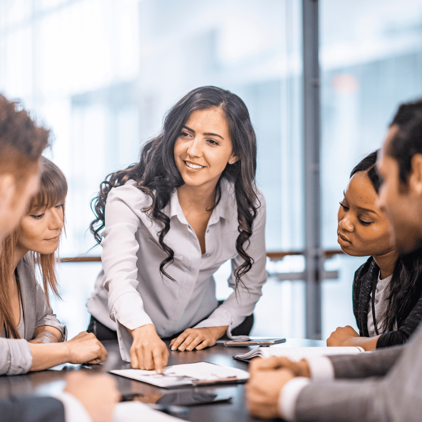 A woman with long curly black hair smiling bending over a table pointing at a document on the table with four people leaning in to look at the document