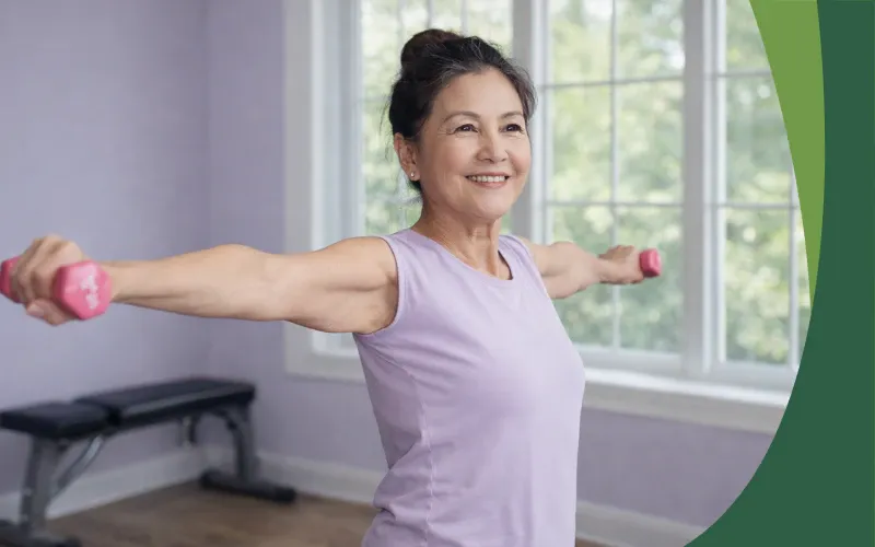 An Asian woman in her 60s with her hair in a bun smiles while strength training at home, holding light dumbbells with her arms extended. She wears a sleeveless lavender top in a bright room with a large window, a light lilac wall, and a weight bench, showing midlife strength and healthy movement.