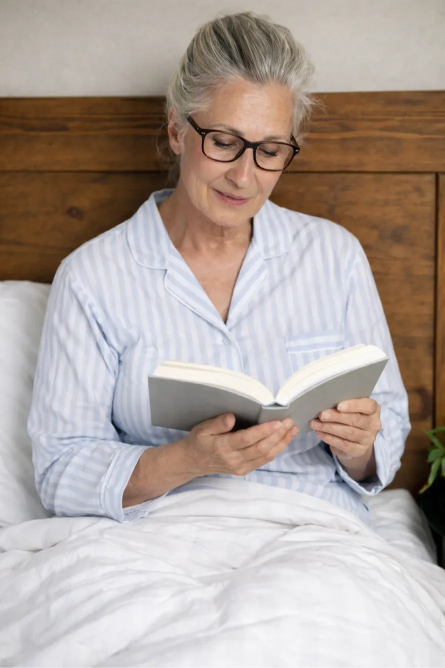 A woman in her 60s with gray hair tied back and wearing glasses sits upright in bed, reading a book held at chest level. She wears light blue striped pajamas and rests against a wooden headboard with white bedding, creating a calm, reflective scene that suggests rest, learning, and midlife brain health.