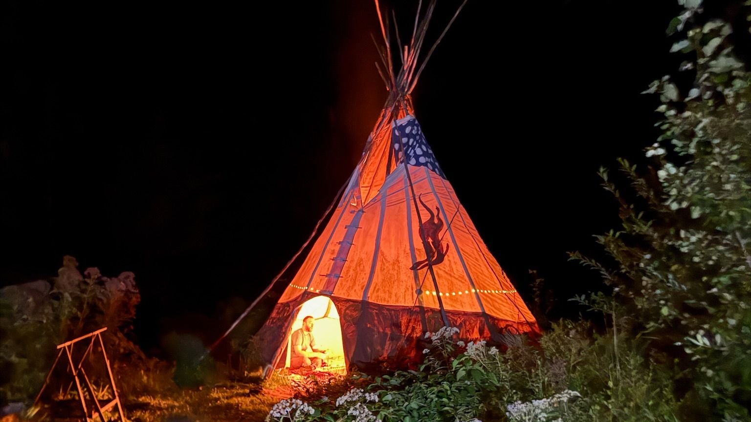 A lit tipi glowing orange at night surrounded by wildflowers during an evening ceremony at a past Metro-Detroit Men's Therapy retreat in Michigan
