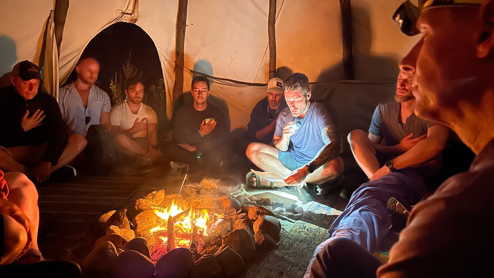 Men sitting in a circle around a fire inside the tipi during an evening council ceremony at a past Metro-Detroit Men's Therapy retreat in Michigan