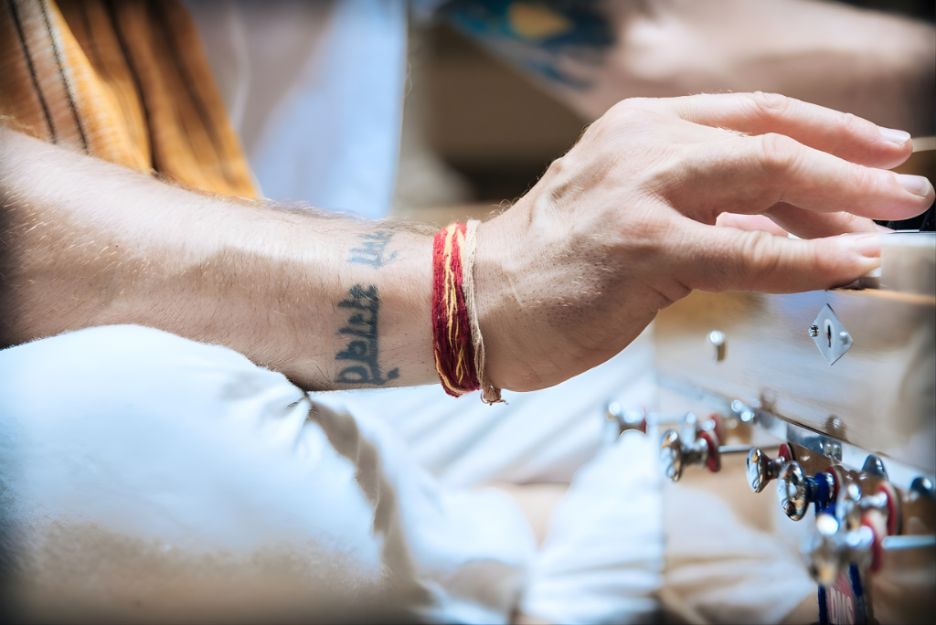 Close-up of a hand playing a harmonium, showing prayer bracelets and a tattoo, during devotional kirtan practice.