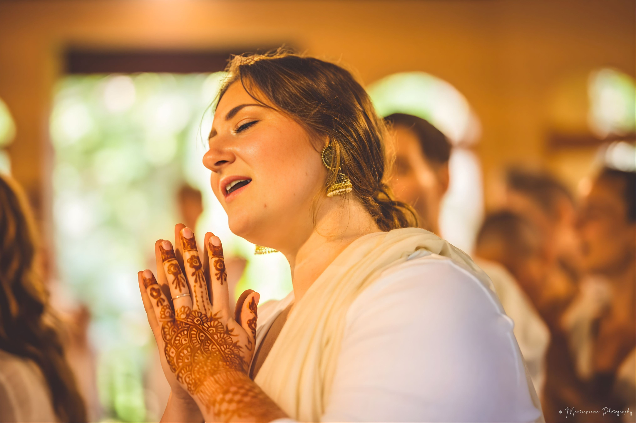Devotee singing during kirtan with hands in prayer, decorated with traditional mehndi, inside a warm devotional gathering space at Govardhan Eco Village in India.