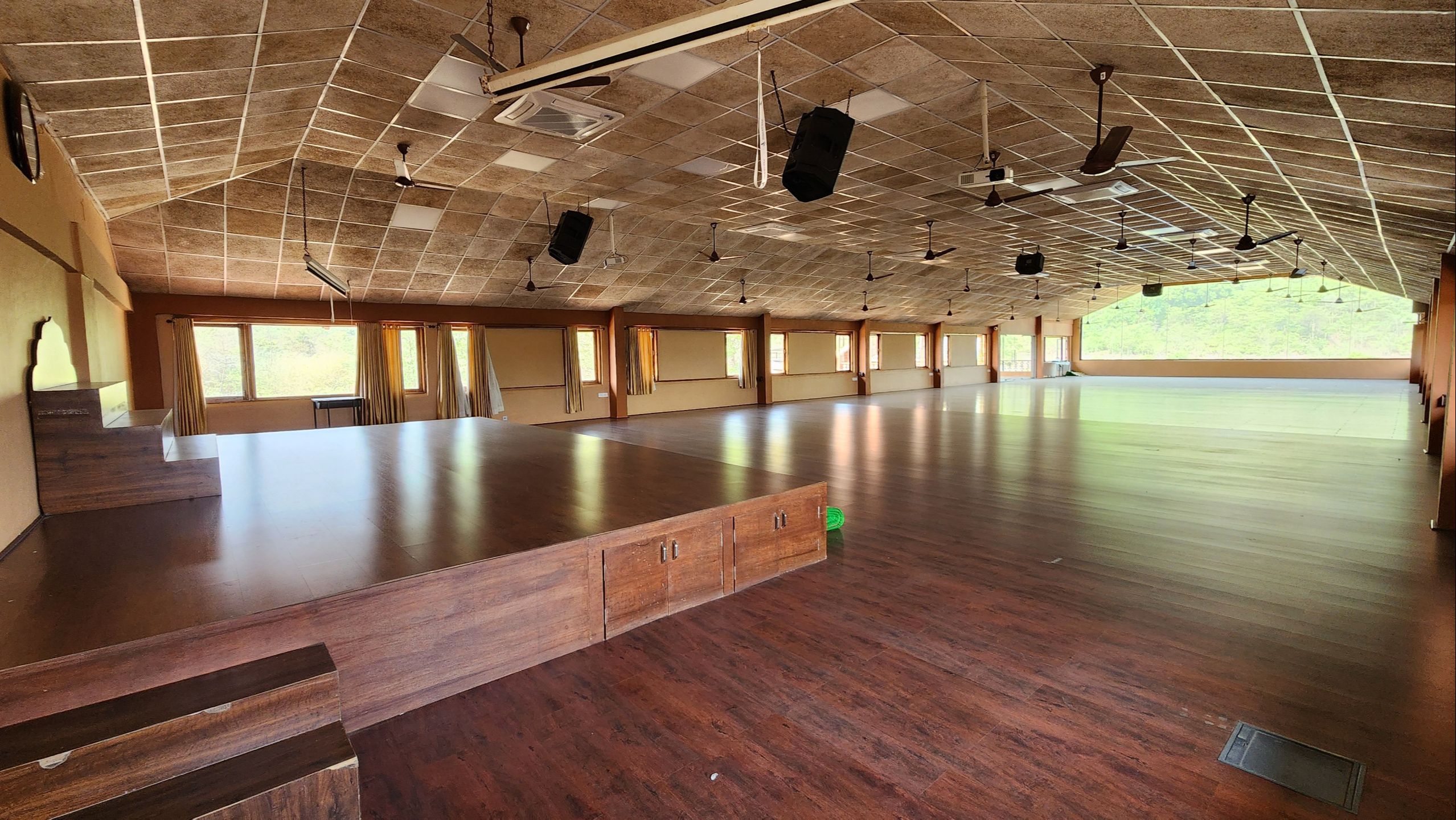 Spacious yoga shala at Govardhan Eco Village in India featuring polished wooden floors, raised teaching platform, natural light from panoramic windows, and serene views of lush greenery—ideal for yoga teacher training and Bhakti Yoga practice.