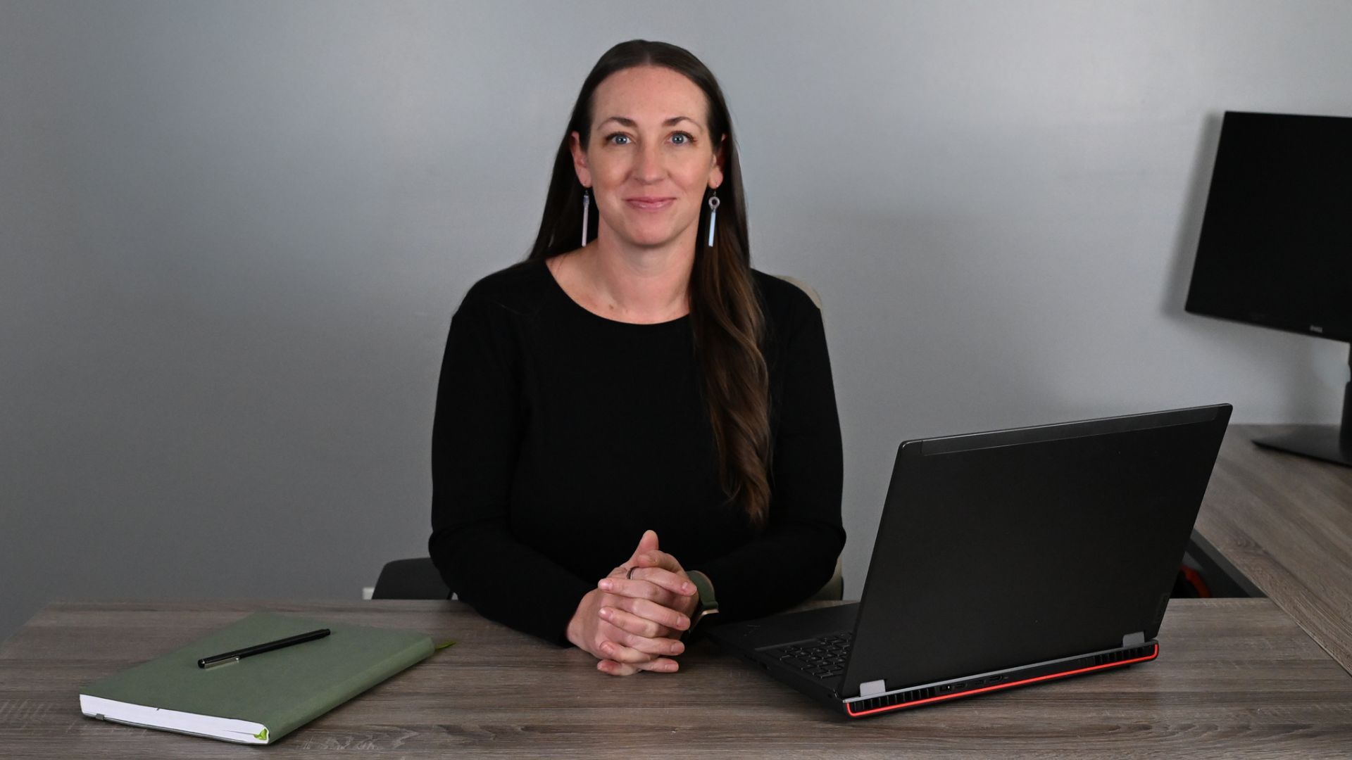 Jill Klott wearing a black shirt sits behind a desk on which is a laptop computer, notebook and pen