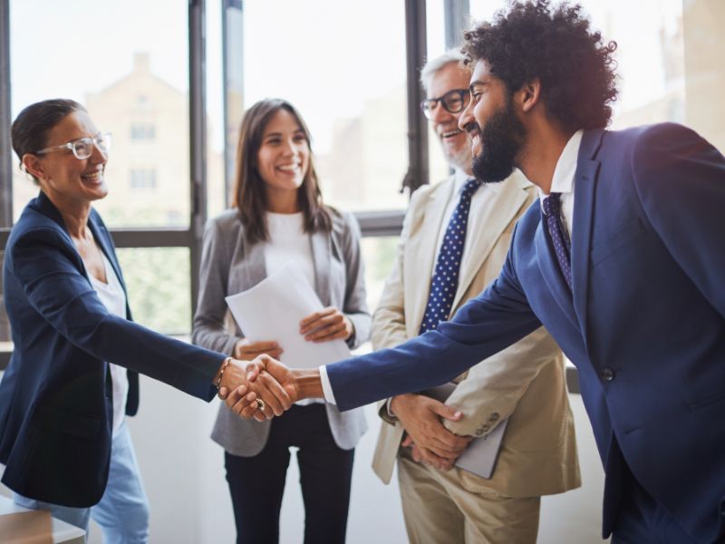 Woman and man in business suits shake hands while two other business people stand and smile