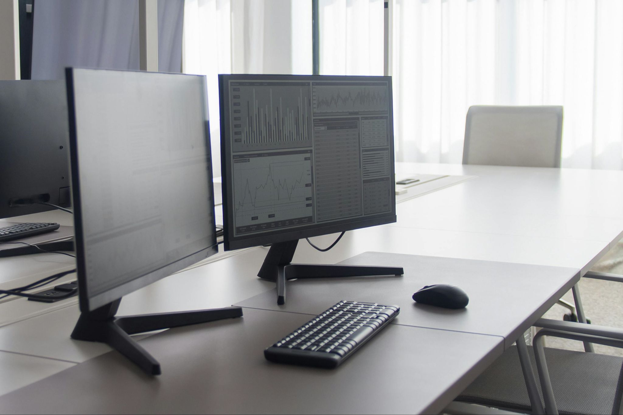 Professional office shows a large conference desk with two computer monitors displaying general spreadsheets and graphs. In the background is softly diffused light from a window and empty chairs.