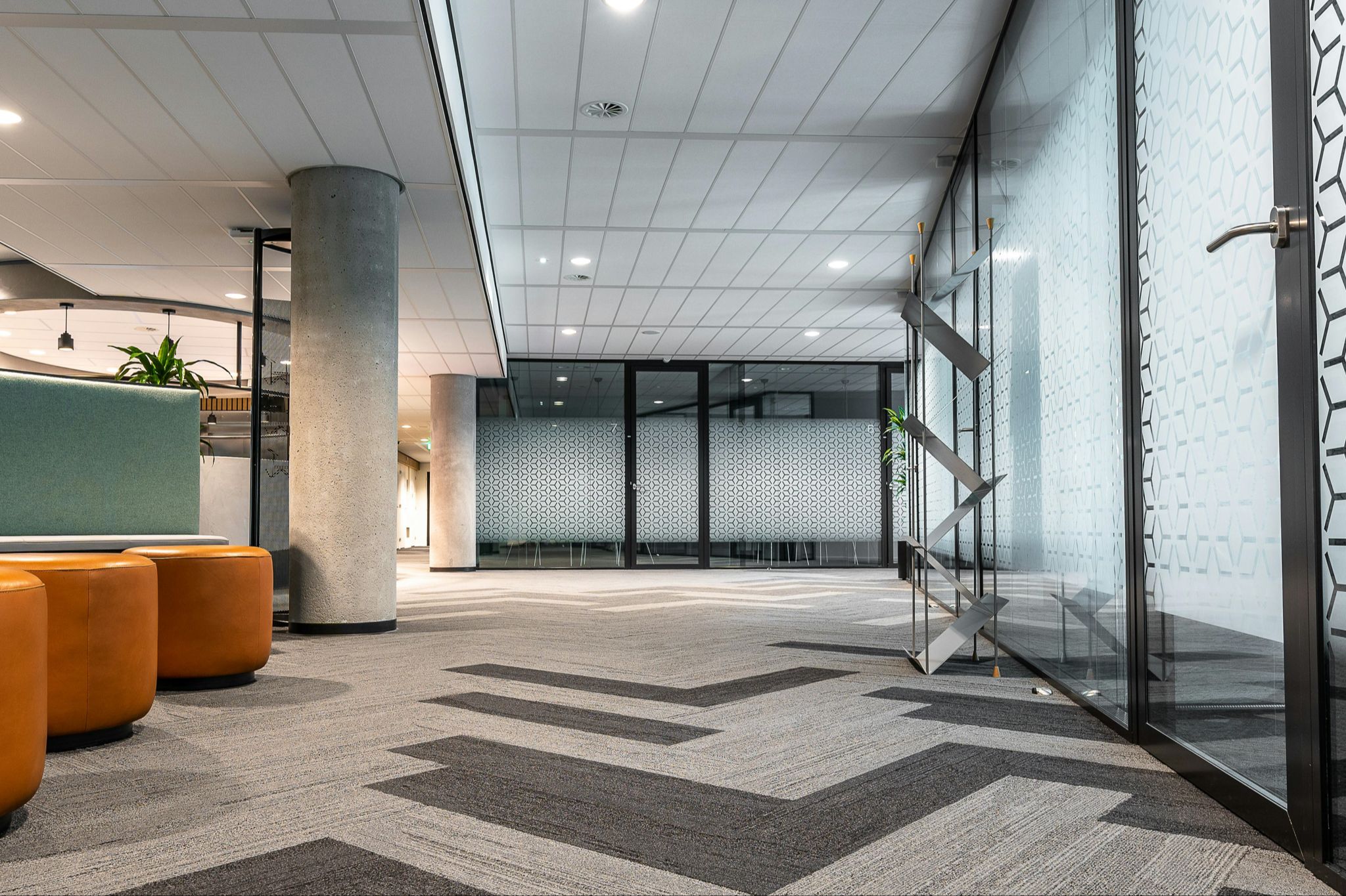 Professional office setting shows two meeting rooms with glass doors and three orange ottomans outside a break area.