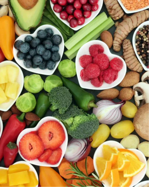 Colorful assortment of whole foods arranged on a table, including berries, citrus, avocado, broccoli, peppers, onions, mushrooms, nuts, and leafy greens. Several fruits sit in heart-shaped bowls, highlighting the connection between real food, nourishment, and heart health.