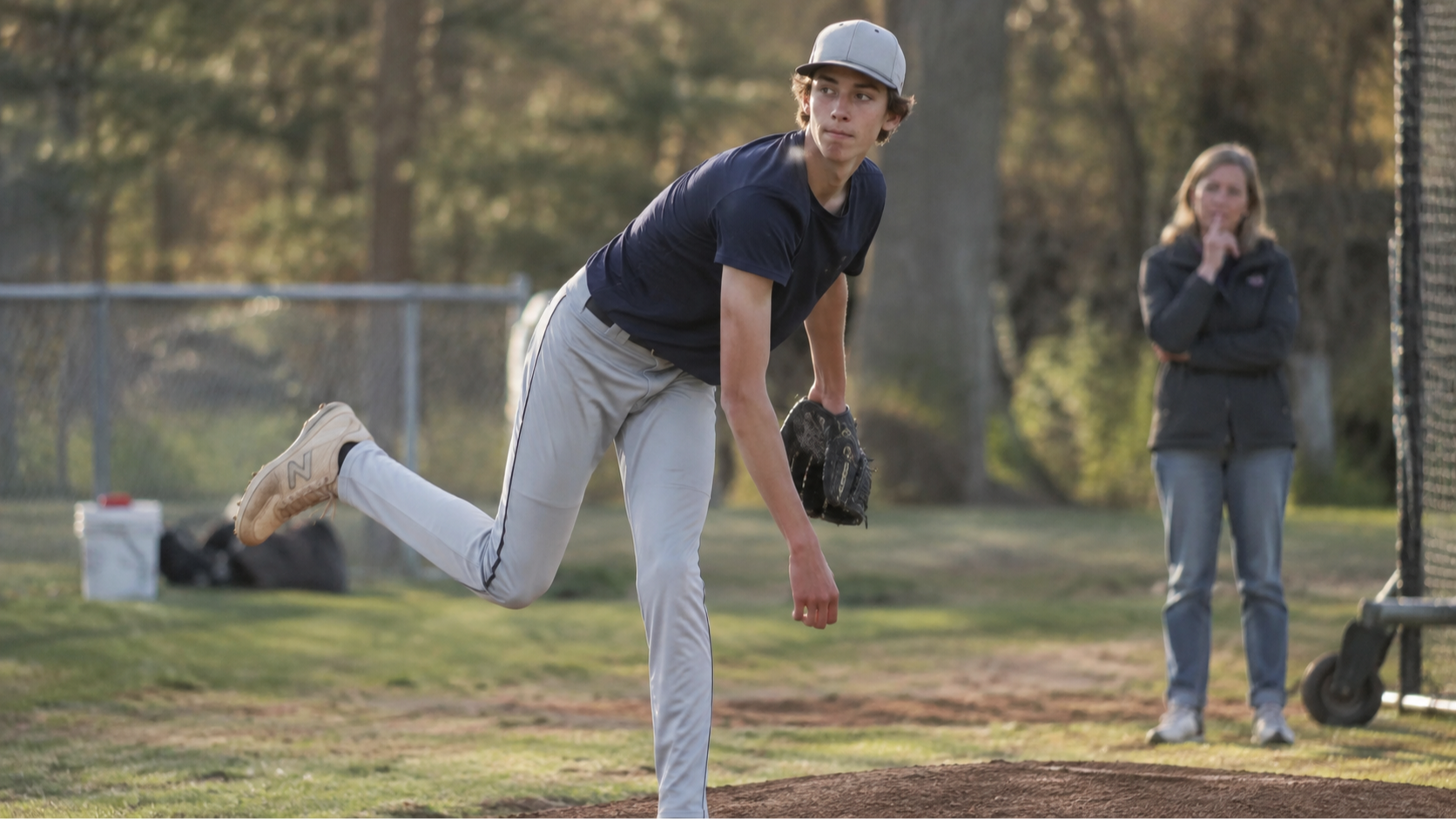 Tall teenage youth pitcher mid-delivery with parent watching from background — youth pitcher growth spurt throwing changes and velocity drop guide for parents