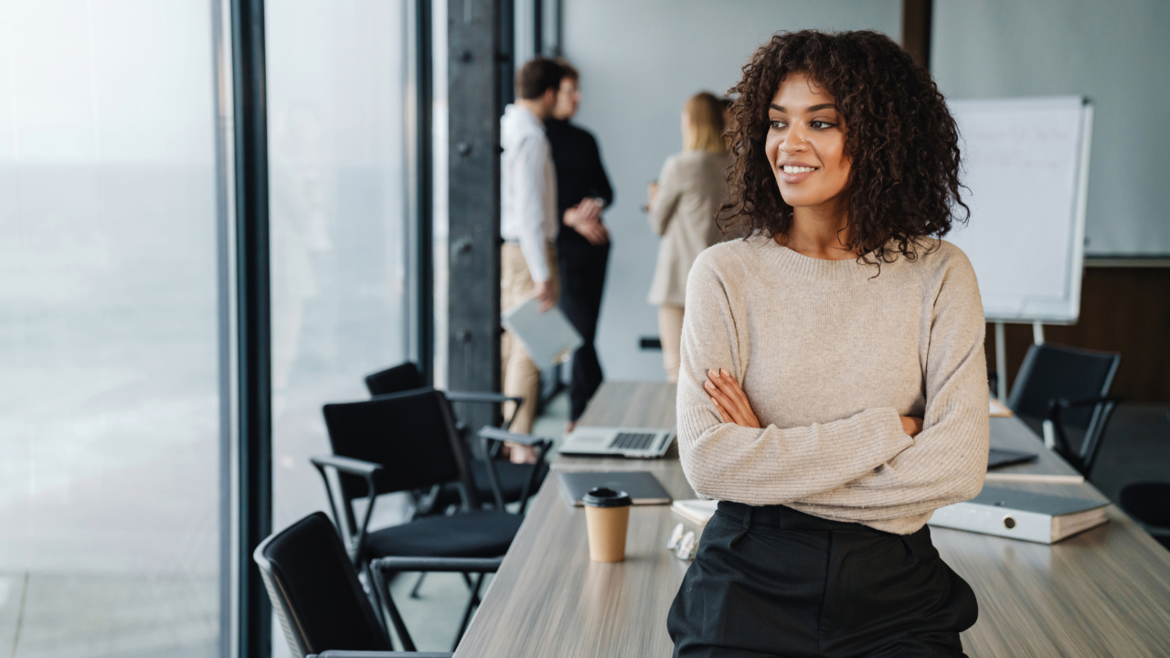 A confident woman with curly brown hair stands in a modern, sunlit office, smiling and looking out a large window with her arms crossed.