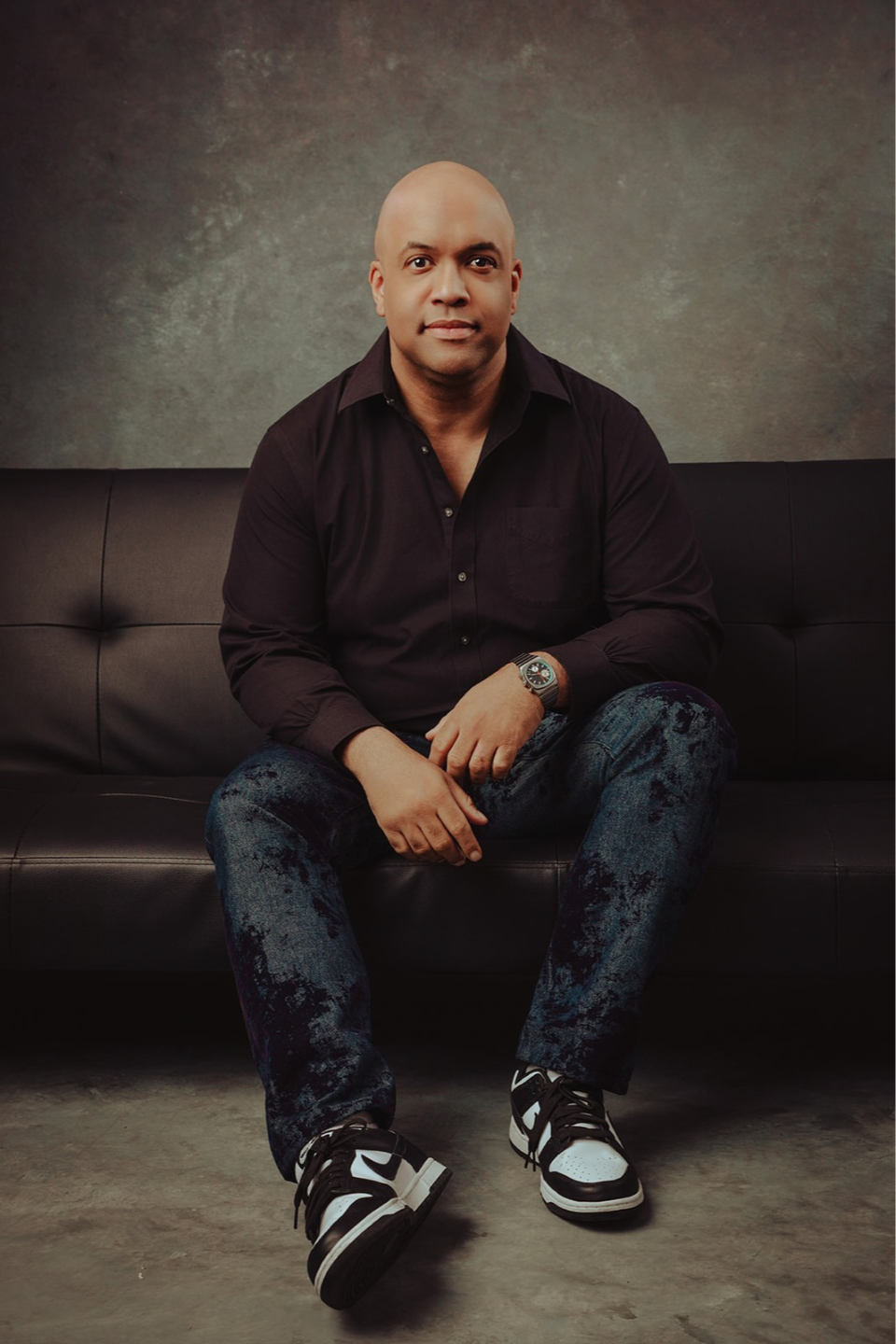 Mike Veny, workplace mental health speaker, author, and Certified Corporate Wellness Specialist®, sits centered on a black tufted leather couch in a professional studio portrait. He leans slightly forward with his elbows resting on his thighs and his hands relaxed, conveying approachability and quiet confidence. He wears a fitted black button-down shirt with the top buttons open, dark blue textured jeans with a subtle distressed pattern, and black-and-white athletic sneakers. A metallic watch is visible on his left wrist.  Mike’s head is clean-shaven, and his facial expression is calm, focused, and present, with direct eye contact toward the camera that suggests trust, authenticity, and leadership. The lighting is soft and even, highlighting his face while creating gentle shadows that add depth and dimension.  The background features a neutral, mottled gray studio backdrop with warm undertones, complementing the overall earthy, grounded color palette. The floor appears to be polished concrete, adding a modern, minimalistic feel. The composition is symmetrical and balanced, with Mike positioned centrally, reinforcing a sense of stability, resilience, and professionalism—qualities aligned with his work in workplace mental health, leadership, and emotional well-being.
