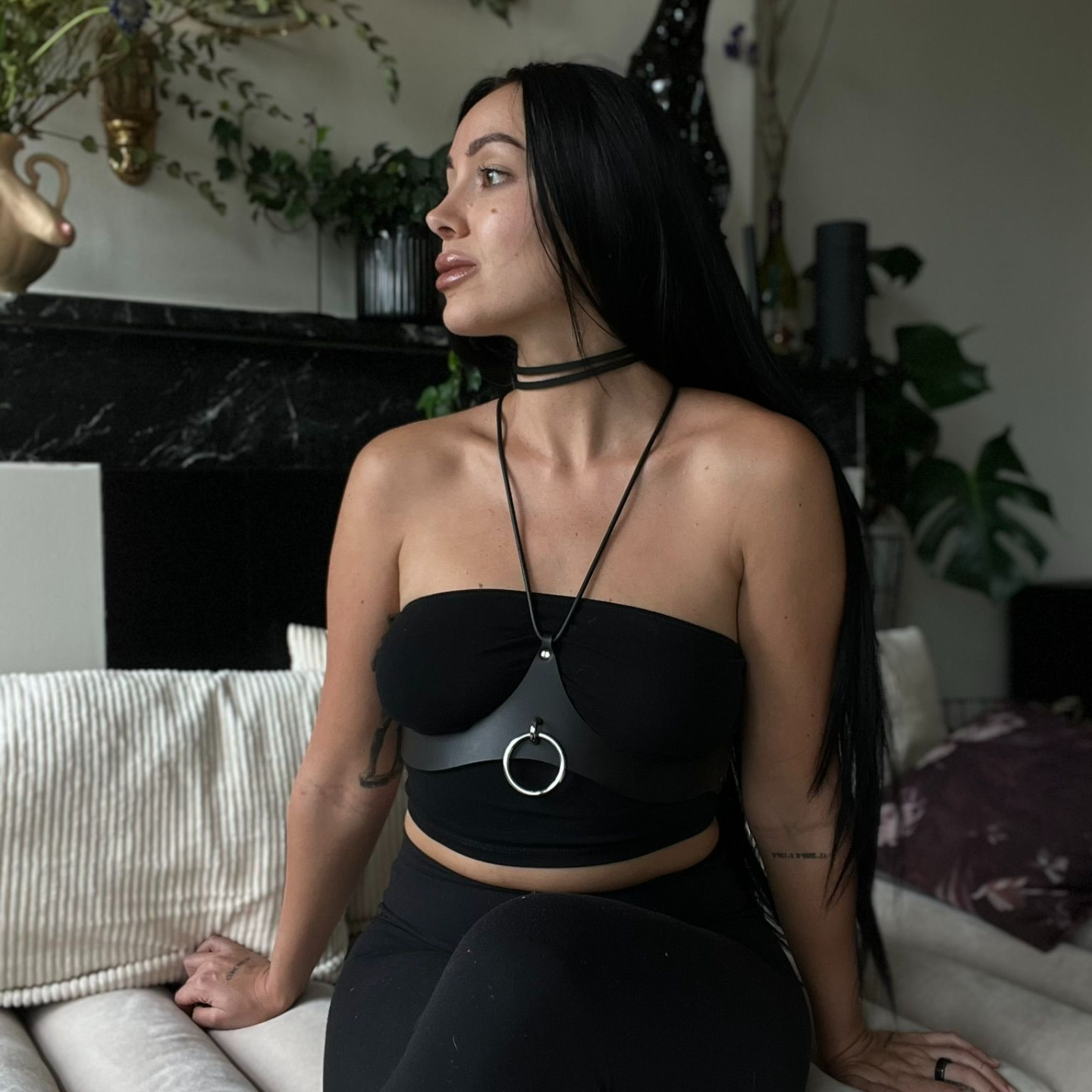 Julieta Chiara Sitting looking sideways wearing all black and a black leather harness and choker