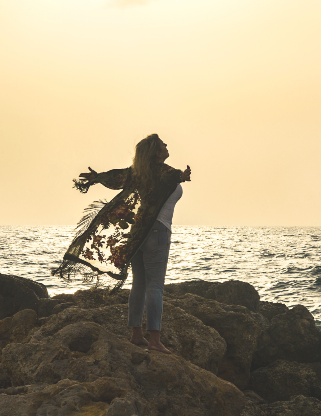 Woman hands wide open on rocks with ocean and sun