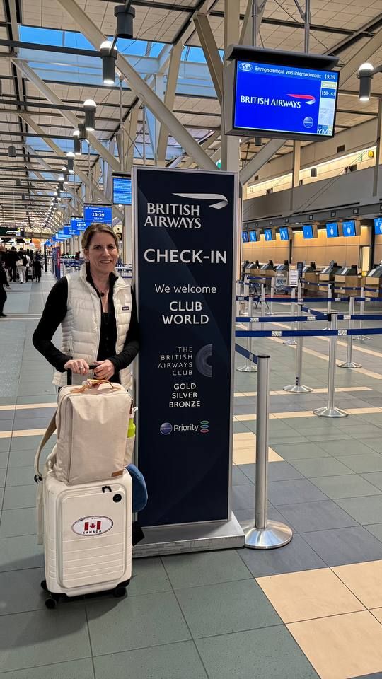 Woman stands smiling with her suitcase beside a British Airways check-in sign in a bright airport terminal.