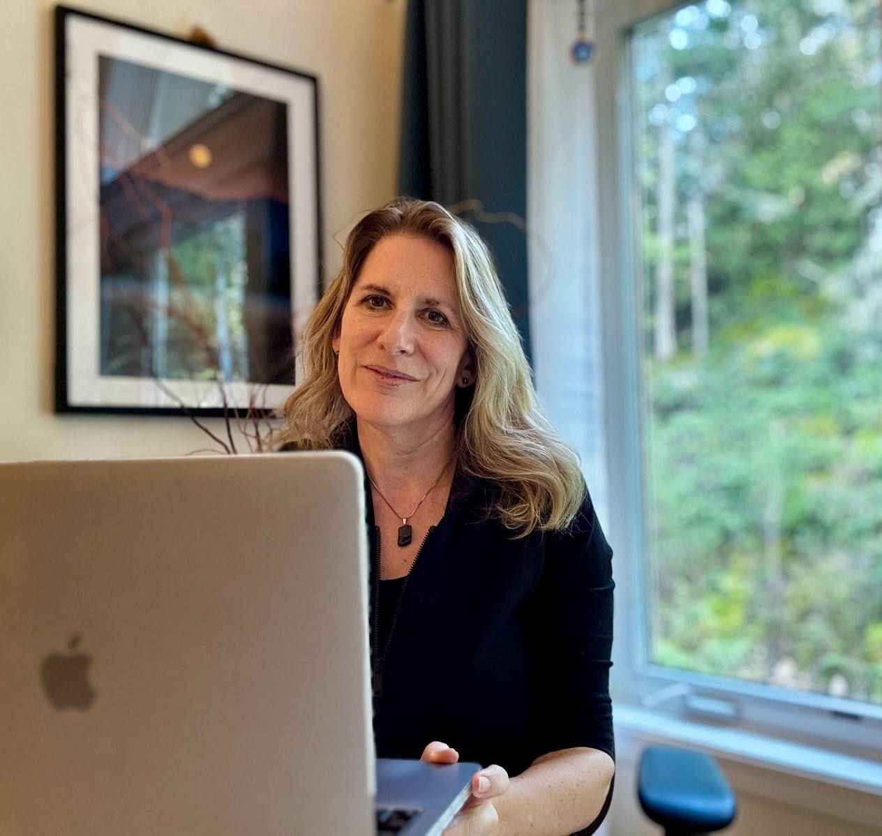 A woman sits at a desk with a laptop, smiling warmly. A large window shows greenery outside, and a framed picture hangs on the wall.