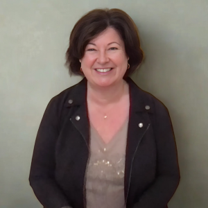 Maria Furlano headshot smiling wearing green and gold shirt with dark grey jacket and soft sage background