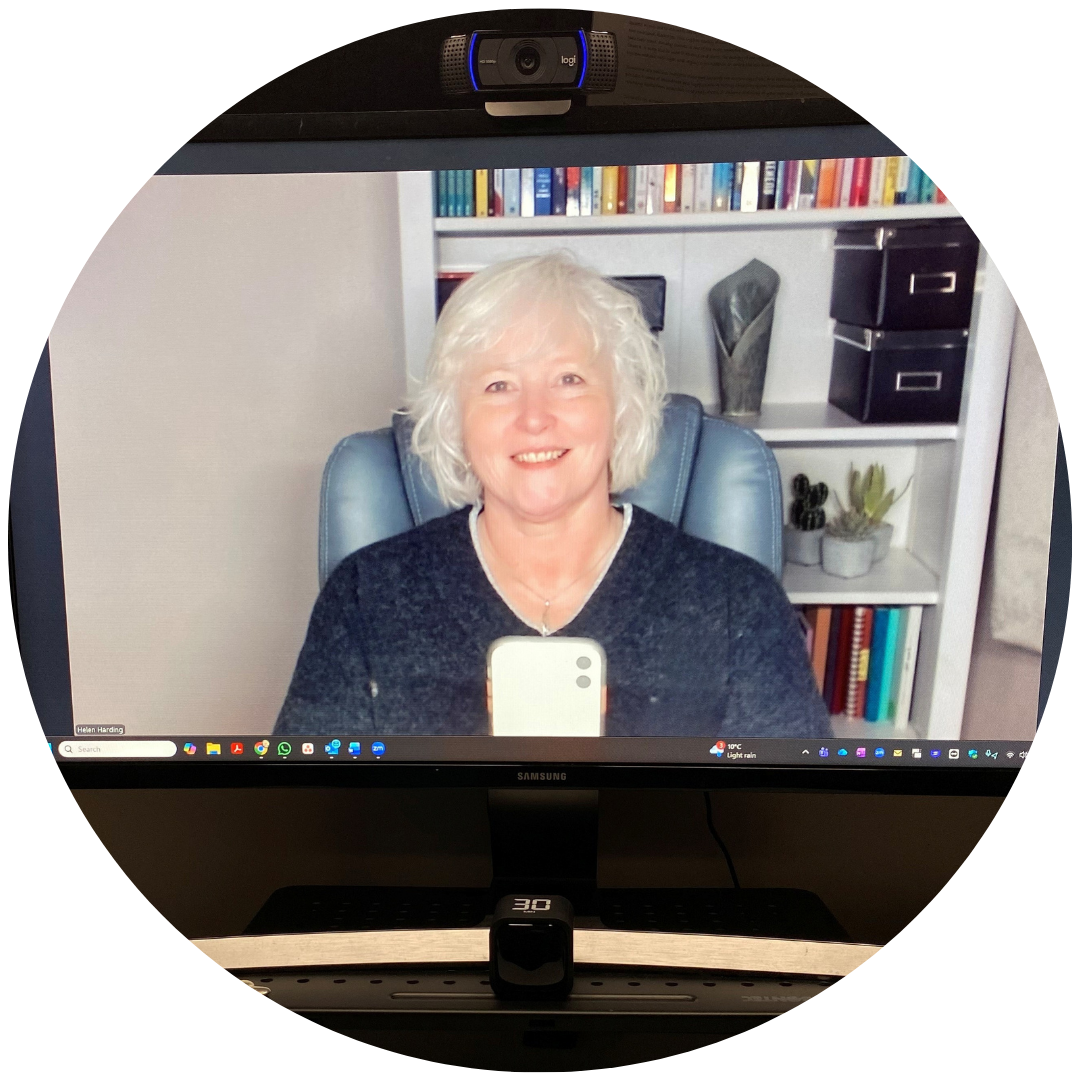 Helen, a 58 year old smiling woman with shortish wavy white hair in a grey jumper taking a selfie of her on a computer screen showing a bookcase in the background.