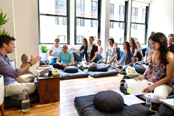 Dr. Miles Neale teaching a meditation class with students seated on cushions in a sunlit studio