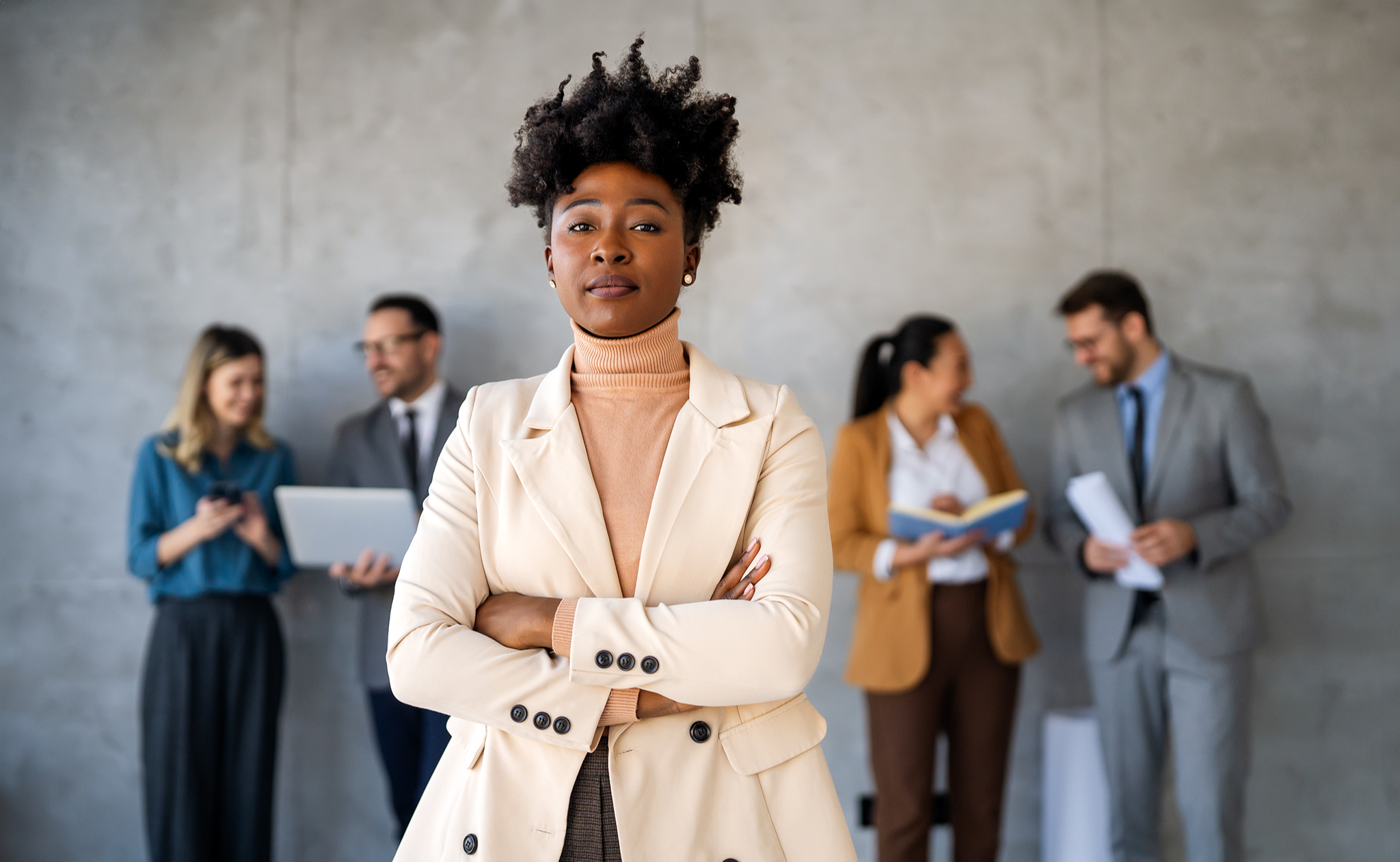 A Black woman in a cream blazer standing with arms crossed, looking directly at the camera with calm authority. Behind her, blurred colleagues converse in pairs. The visual embodiment of taking up space — present in the room, settled in herself, not shrinking or performing.