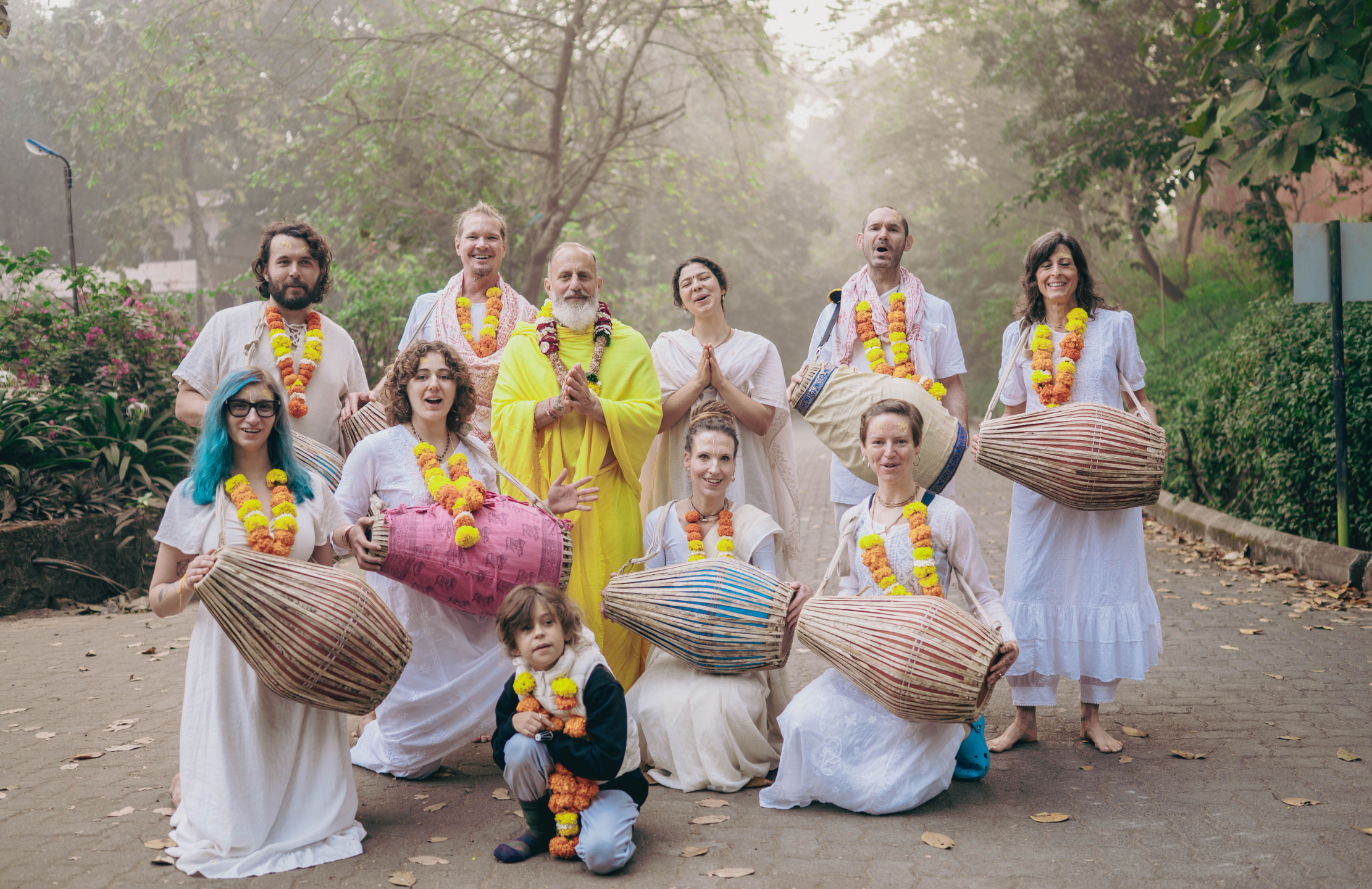 Mrdanga School graduation ceremony at Govardhan Eco Village in India featuring students and teachers in traditional attire playing mrdanga drums, wearing marigold garlands, and celebrating with kirtan in a peaceful outdoor setting.