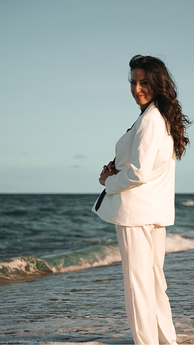 Woman in a white suit sits on a wooden chair by the sea, smiling with her eyes closed