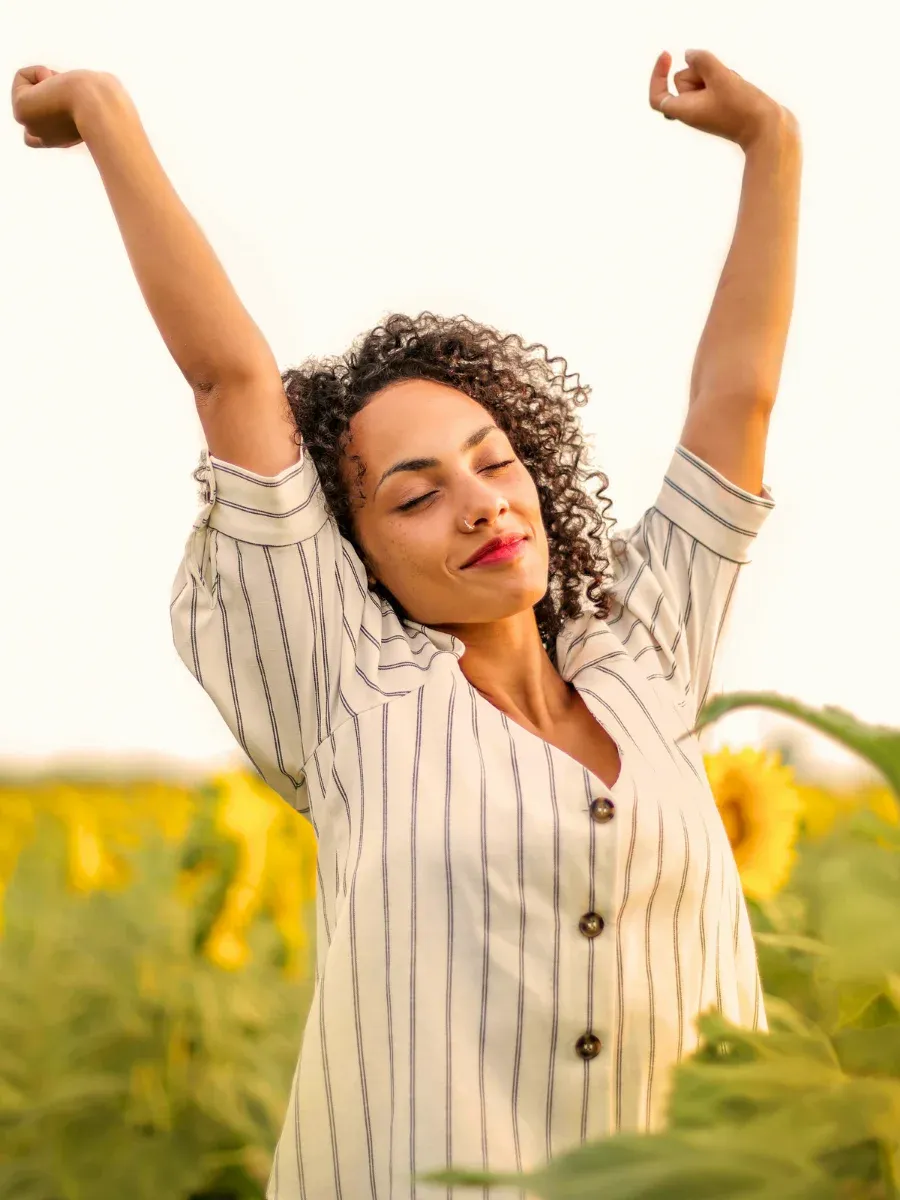 Woman with arms raised joyfully in a sunflower field representing self-nurturing and coming back to yourself