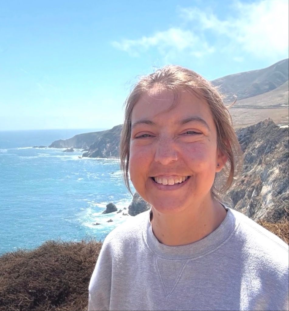 a woman is smiling in front of the ocean with a rocky shore behind her.