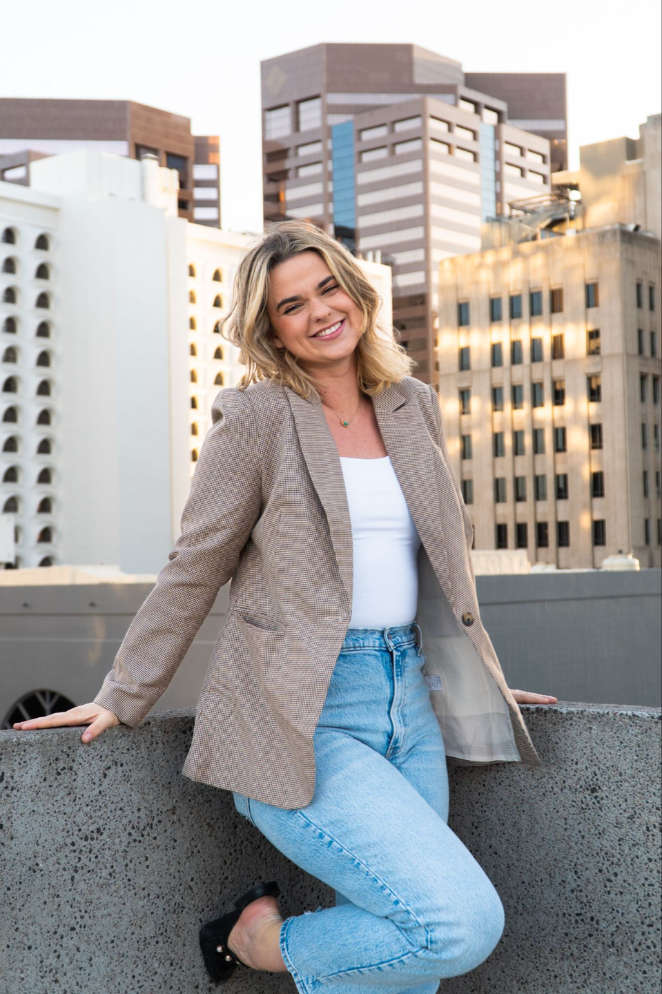 a woman stands in business-casual attire on a rooftop surrounded by buildings in the city.