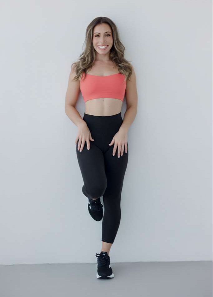 Jennah Jacobs smiling and leaning against a blank grey wall, one foot on the wall behind her, wearing sports bra and tights.