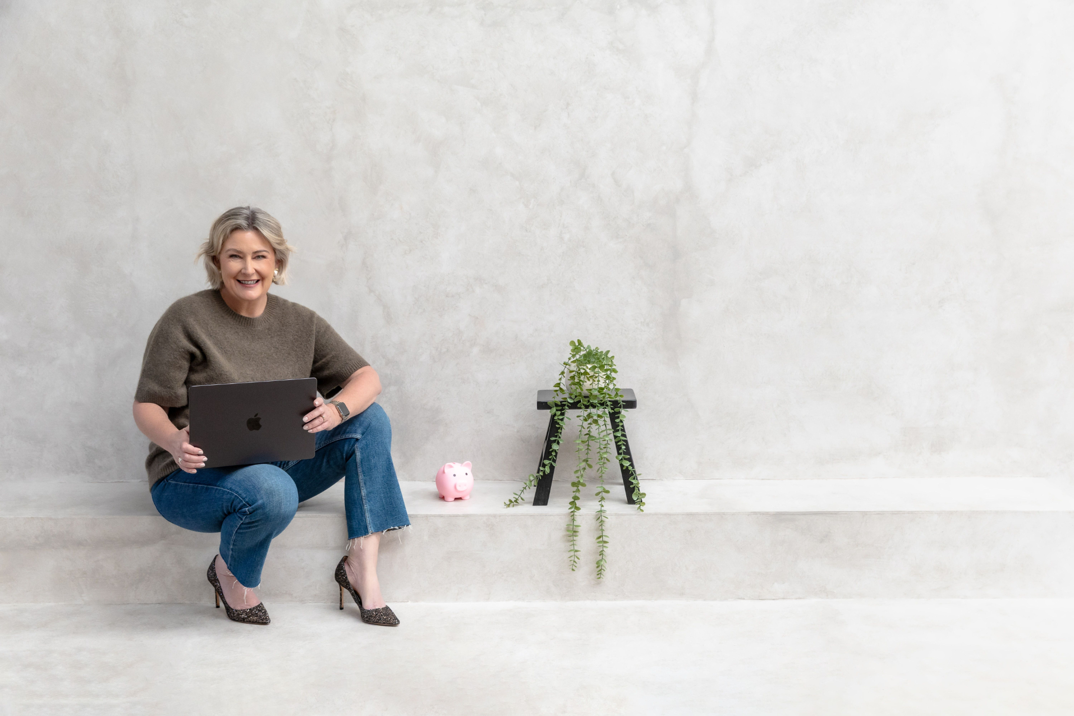 An image of Kylie Sultana, Money Coach and founder of The Money Brew, sitting elegantly in a soft white bouclé armchair, laughing warmly as she works on her silver laptop. She has chic short blonde hair and wears a tasteful olive-green knit top with a delicate floral brooch, relaxed light-wash jeans, and stylish nude pointed heels. Behind her, a sheer curtain and a sculptural black floor lamp create a calm, modern Australian home studio setting, reflecting the warm, judgement-free coffee-chat energy of The Money Brew personal finance education for women.