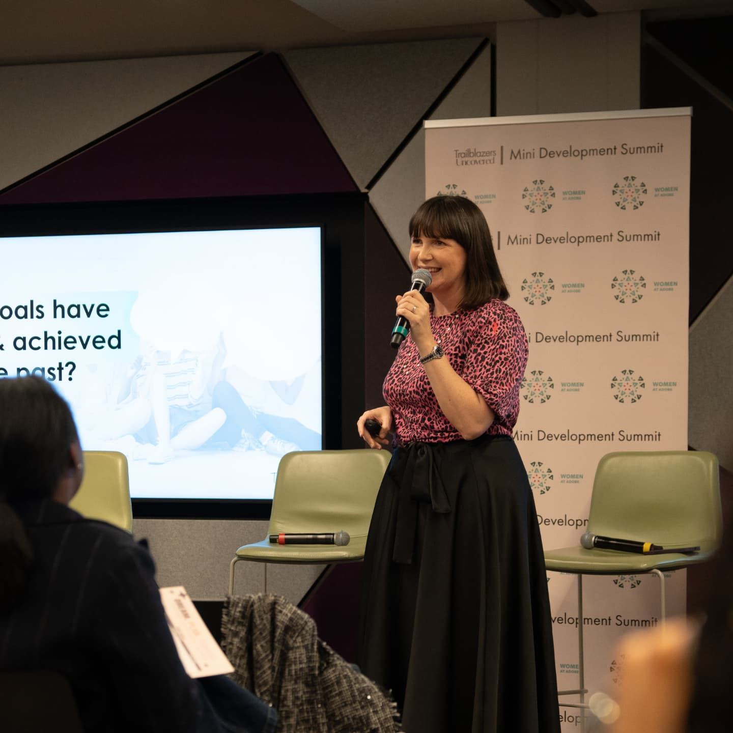 Tara Rule speaking on stage at a leadership development event, holding a microphone and presenting to an audience, with presentation slides and event branding visible in the background.